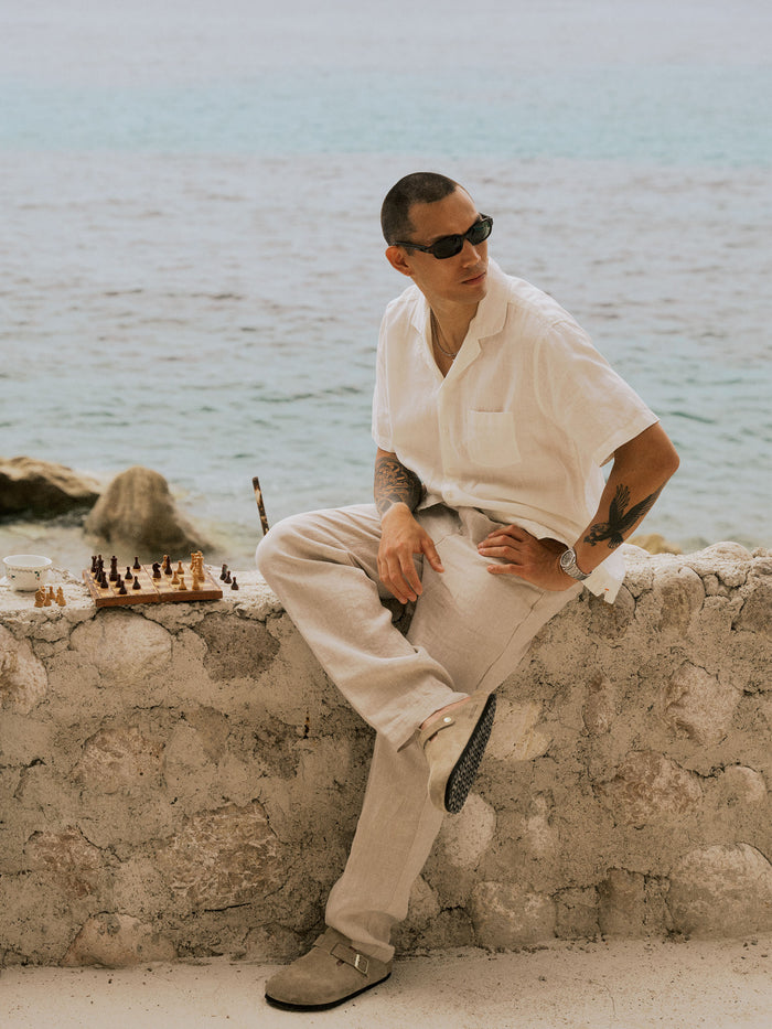 The Holiday Linen Trouser in Natural — sitting on a stone wall next to a chessboard