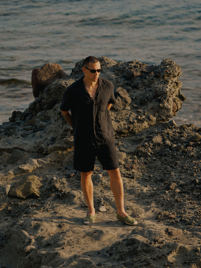 The Holiday Linen SS Shirt in black — worn on a rocky beach by the ocean