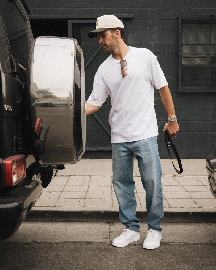 A man stands outdoors near the back of a black SUV wearing a white t-shirt, a cap, and The Jeans Relaxed Straight Fit in Light Midwash by c'est normal.