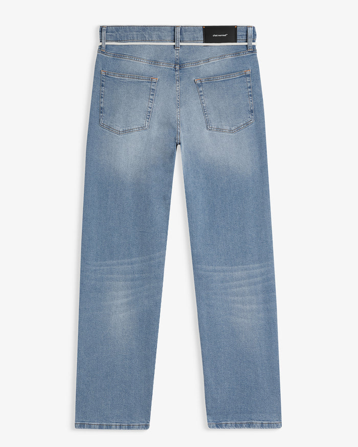A back view of The Jeans Relaxed Straight Fit in Light Midwash, laid flat on a white background. The image highlights the two back pockets, the subtle orange accent stitching, and the rubber patch at the back waist with the c'est normal logo.