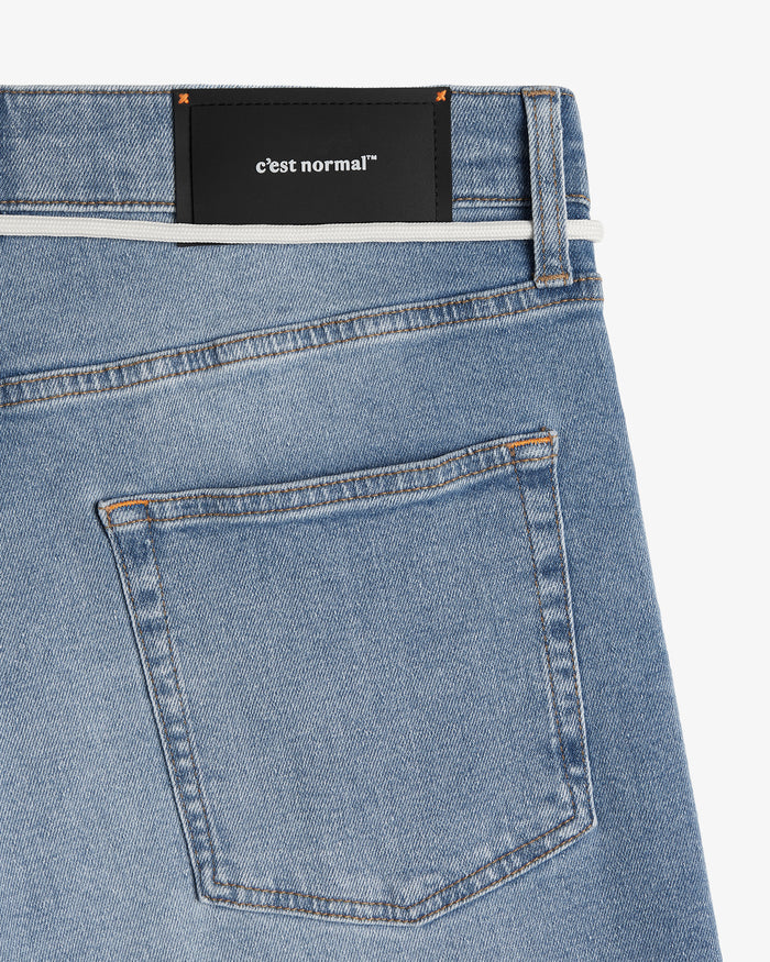 A close-up shot of the back of The Jeans Relaxed Straight Fit in Light Midwash. The image highlights the right back pocket, subtle orange accent stitching, and the black rubber patch with the white c'est normal logo at the back waist.