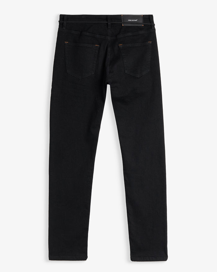 A back view of The Jeans Skinny Fit in Jet Black, laid flat on a white background. The image highlights the two back pockets, subtle orange accent stitching, and the black rubber patch with the white c'est normal logo at the back waist.