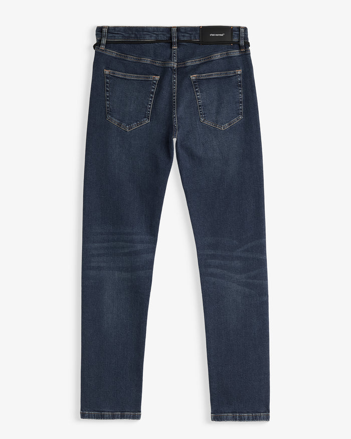 A back view of The Jeans Skinny Fit in Midwash, laid flat on a white background. The image highlights the two back pockets and the black rubber patch with the c'est normal logo at the waist.