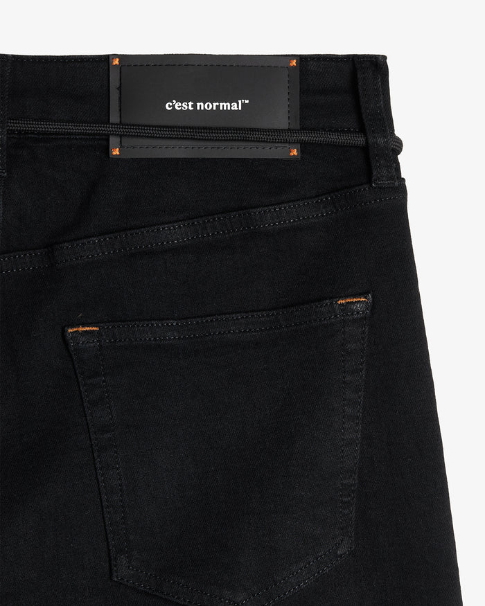 A close-up shot of the back waist of The Jeans Skinny Fit in Jet Black, laid flat on a white background. The image highlights the black rubber patch with the c'est normal logo, back pocket, and subtle orange accent stitching.