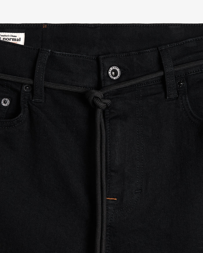 A detailed close-up of the front waistband of The Jeans Skinny Fit in Jet Black, focusing on the branded button, black drawstring, and subtle orange accent stitching. A label on the inside features the c'est normal logo.