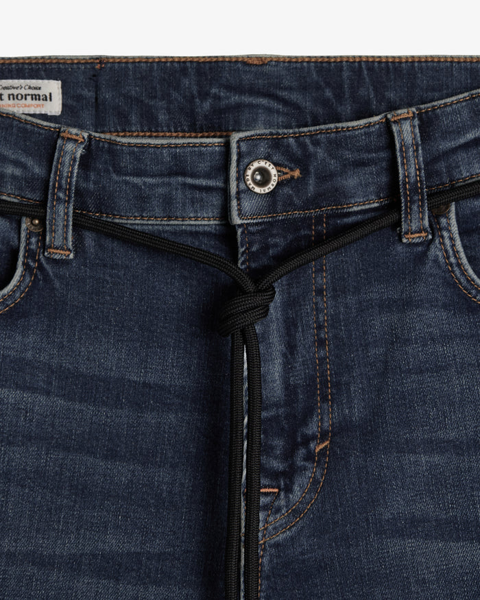 A close-up shot of the front waist area of The Jeans Skinny Fit in Midwash, highlighting the metal button, subtle orange accent stitching, and a black drawstring detail. A small white label with the c'est normal logo is visible on the inner waistband.