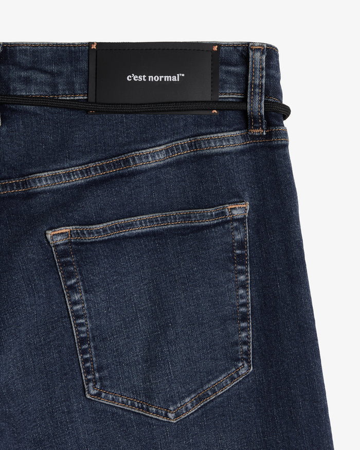 A detailed close-up of the back waist and pocket of The Jeans Slim Fit in midwash. The image highlights the black rubber patch with the white c'est normal logo, subtle orange accent stitching, and a black cord running through the belt loops.