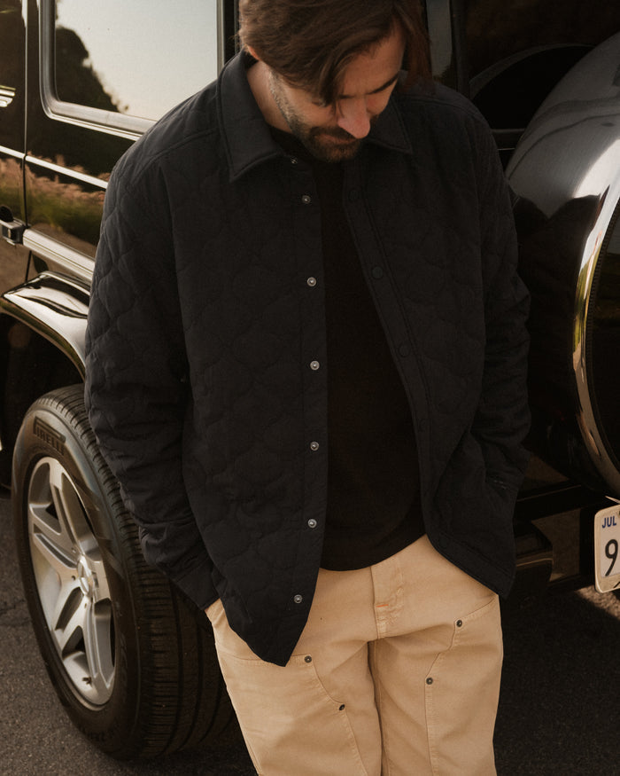 A man looking down while wearing The Quilted Overshirt in black over a black t-shirt and tan pants. He is standing in front of a black vehicle, with his hands in his pockets.