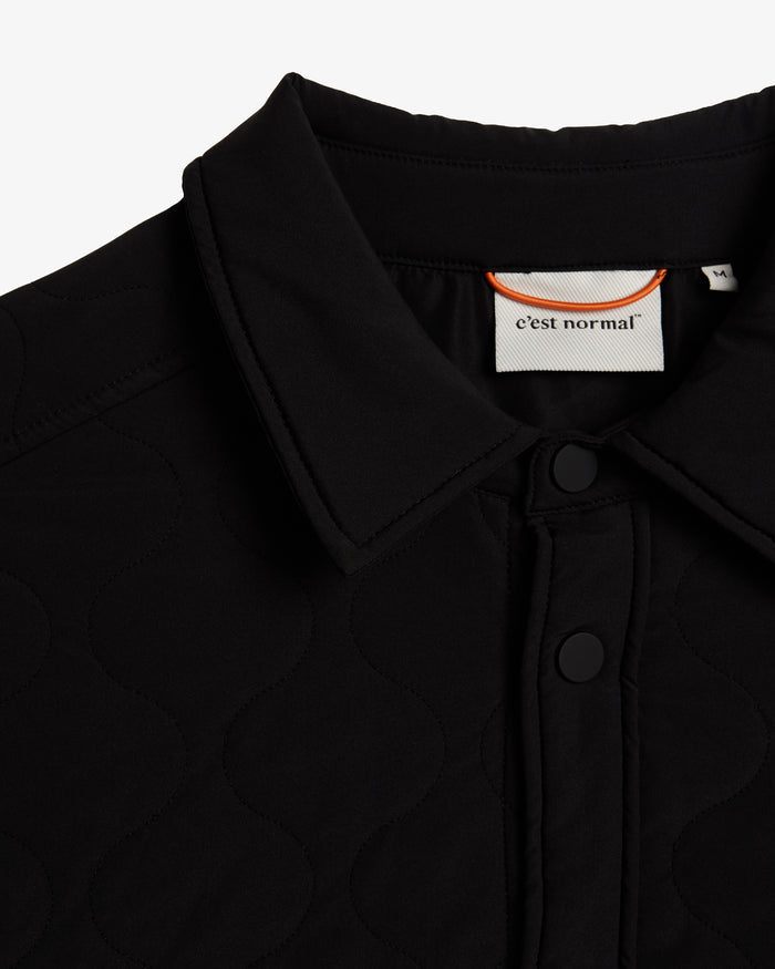 A close-up detail of The Quilted Overshirt in black, focusing on the collar, front snap buttons, and the signature wave quilting. The image highlights the inside neck tag featuring the c'est normal logo and an orange hanging loop.