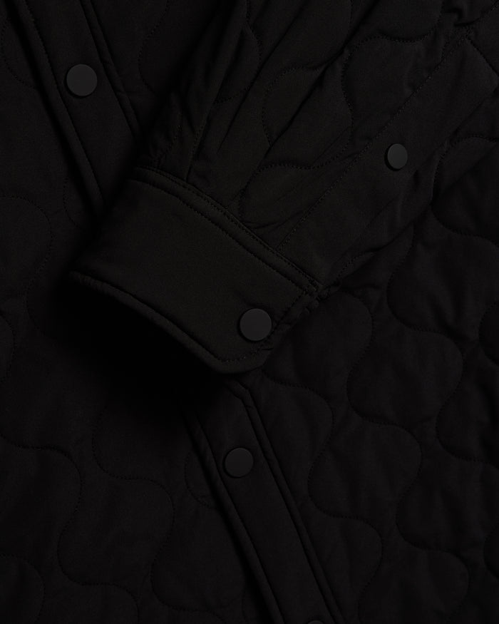 A close-up shot of The Quilted Overshirt in black, highlighting the signature wave quilting pattern and the matte black snap buttons on the cuff and front placket.