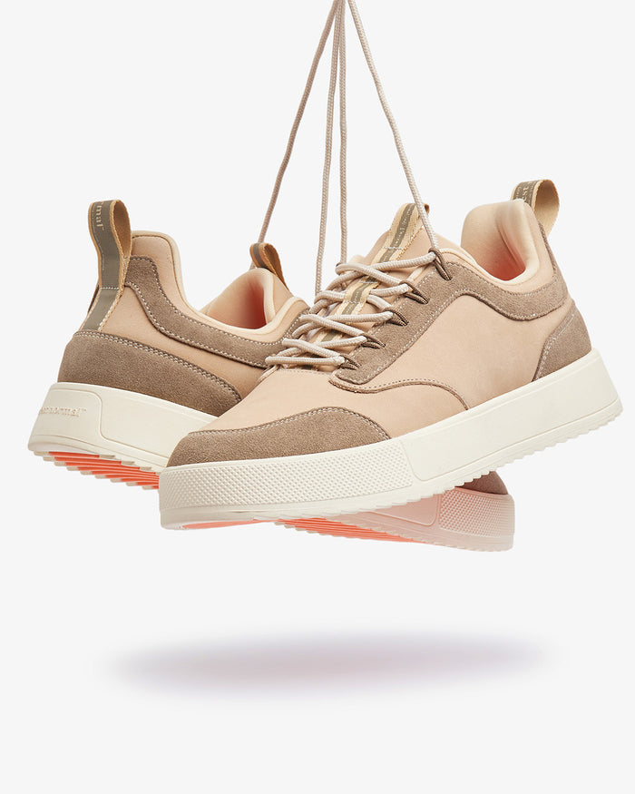 A pair of The Sneakers 2.0 Leather in sand leather, suspended by their laces against a light background. The image highlights the tone-on-tone leather and suede upper, the white midsole, and the c'est normal logo on the heel.