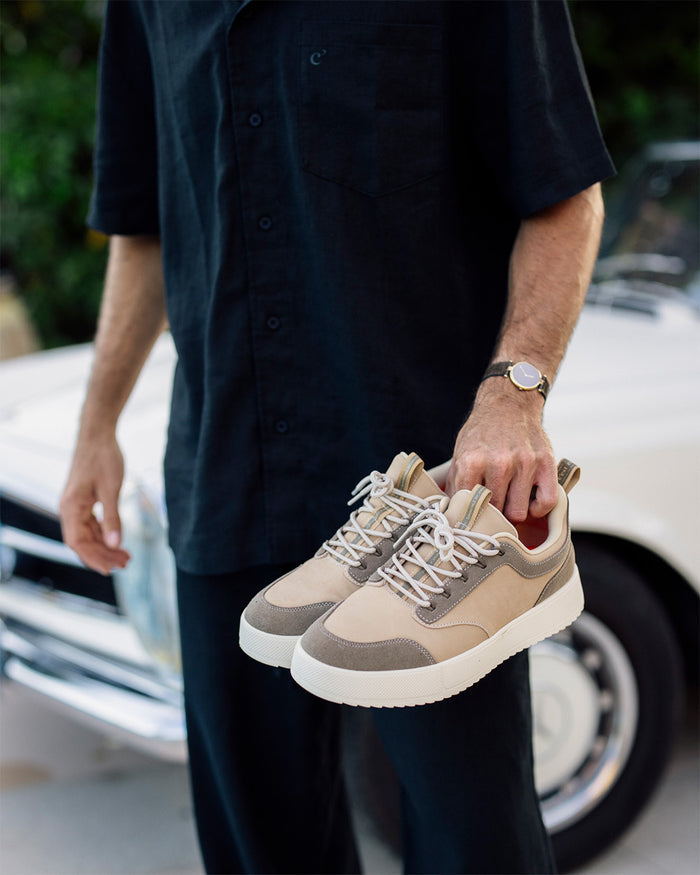 A man holding a pair of The Sneakers 2.0 Leather in sand leather, standing in front of a white car. The image highlights the tone-on-tone leather and suede upper, and he is wearing a black short-sleeved shirt with a subtle c'est normal logo.