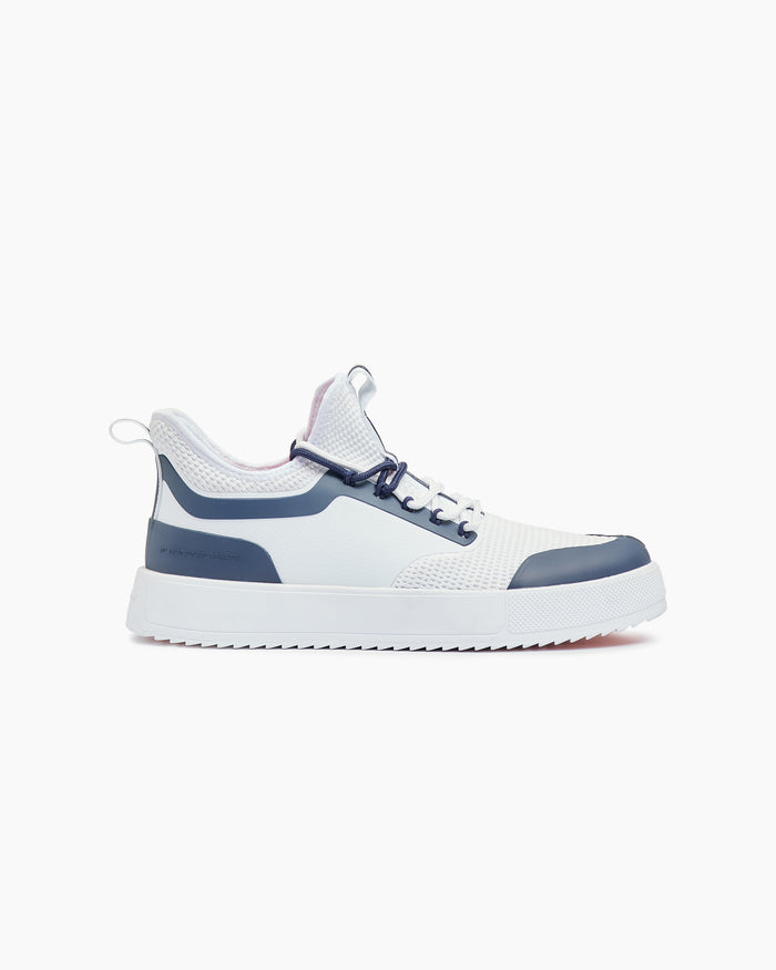 A side view of The Sneakers 2.0 in white and navy, laid flat on a white background. The image highlights the white mesh upper, navy rubber accents, and the chunky white sole of the c'est normal sneaker.