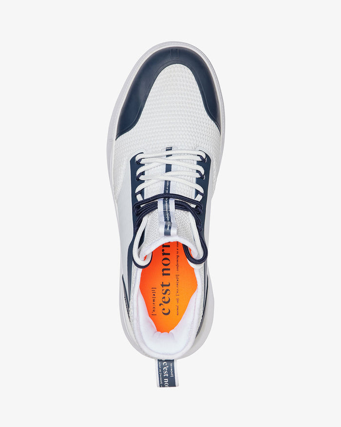 A top-down view of The Sneakers 2.0 in White/Navy, laid flat on a white background. The image highlights the white mesh upper, navy blue accents, custom lace loops, and the original orange insole featuring the c'est normal logo.