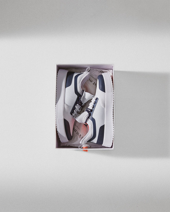 A pair of The Sneakers 2.0 in white and navy, laid inside an open white shoe box lined with c'est normal branded tissue paper, set against a plain light background.
