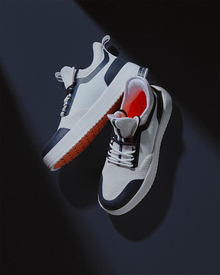 A pair of The Sneakers 2.0 in white and navy, placed on a dark background. The image highlights the white mesh upper with navy accents, the original orange rubber grid sole, and the c'est normal logo tag on the heel.