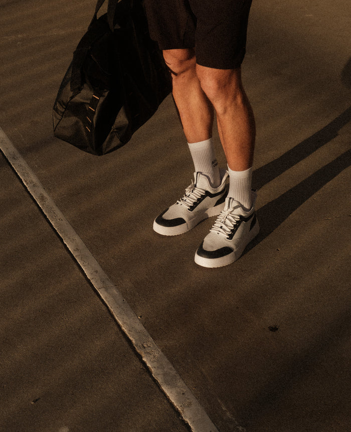 A person wearing The Sneakers 2.0 in white and navy, along with white c'est normal socks, holding a black gym bag while standing on an outdoor court.