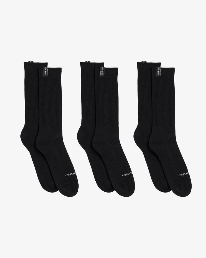 Three pairs of The Sock in black, laid flat on a white background. The image highlights the mid-calf length and the c'est normal logo on the cuff tags and across the toes.