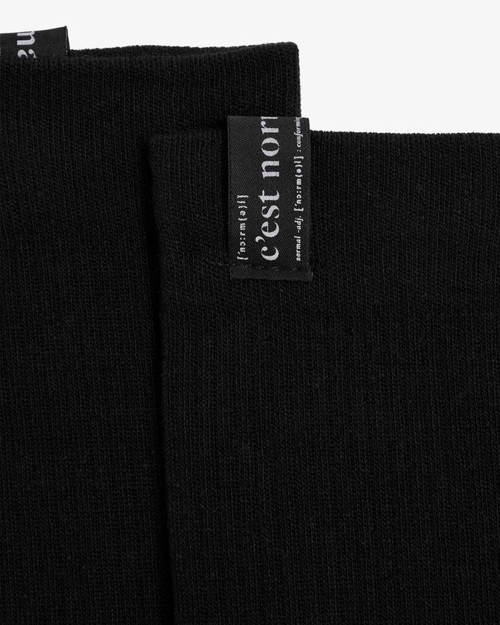 A close-up of The Sock in black, focusing on the cuff tag that features the white c'est normal logo.