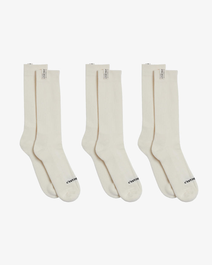 Three pairs of The Sock in cream, laid flat side-by-side on a white background. The image highlights the ribbed mid-calf design and the c'est normal logo on both the cuff tag and across the toe.