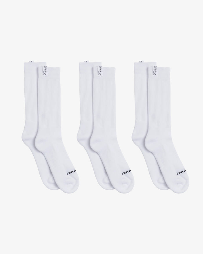A front view of The Sock White 3-Pack, laid flat on a white background. The image highlights the three pairs of white mid-calf length socks, featuring the c'est normal logo on the cuff tag and across the toe.
