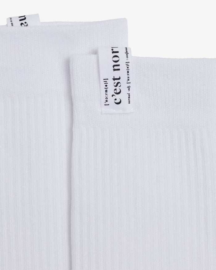 A detailed close-up of The Sock in white, focusing on the ribbed fabric and the cuff tag featuring the black c'est normal logo and definition.