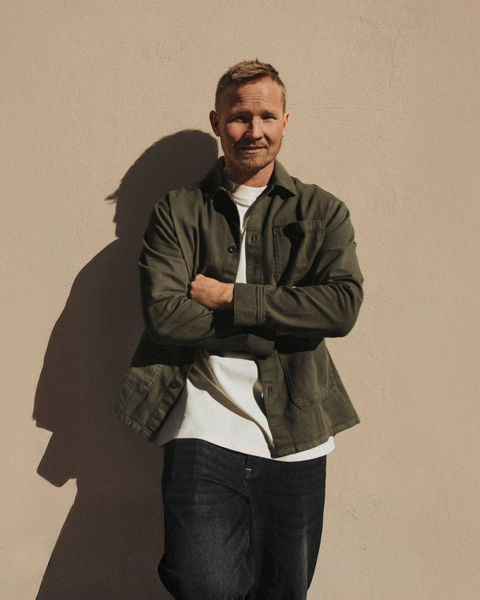 A man leaning against a beige wall with his arms crossed, wearing The Workwear Overshirt in olive green layered over a white t-shirt and dark jeans.