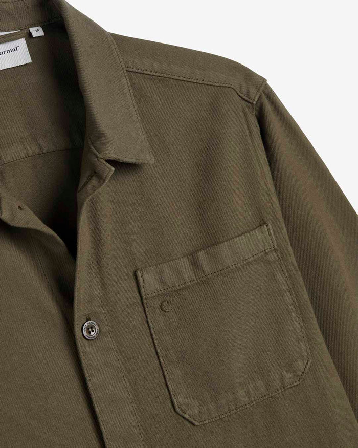 A detailed close-up of The Workwear Overshirt in olive green, laid flat on a white background, highlighting the collar, the left chest patch pocket with a tone-on-tone embroidered c'est normal logo, and a branded button.