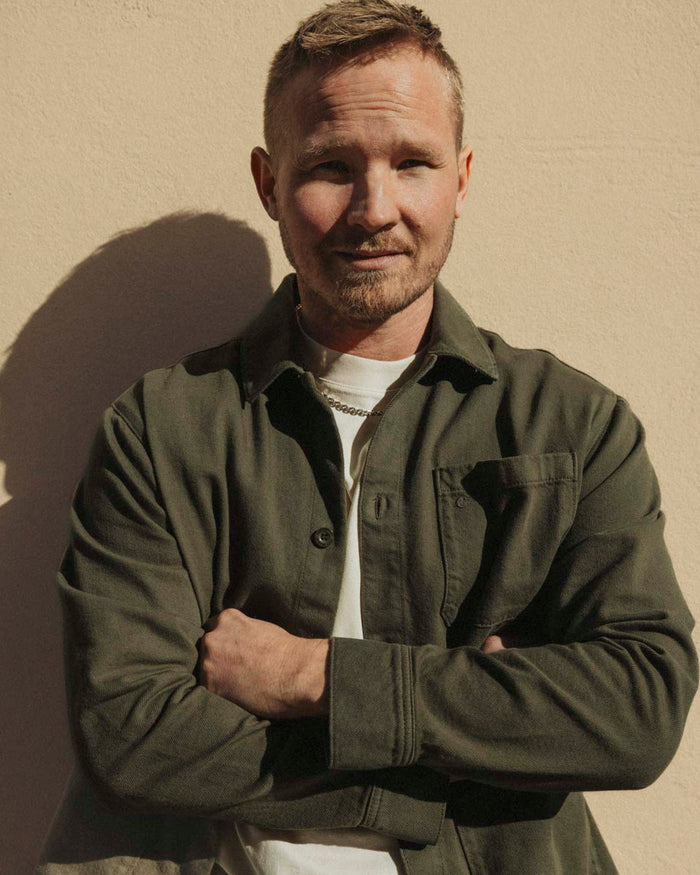 A man stands with his arms crossed, wearing The Workwear Overshirt in Olive Green over a white t-shirt. The image highlights the shirt's patch pocket on the left chest and its straight fit.
