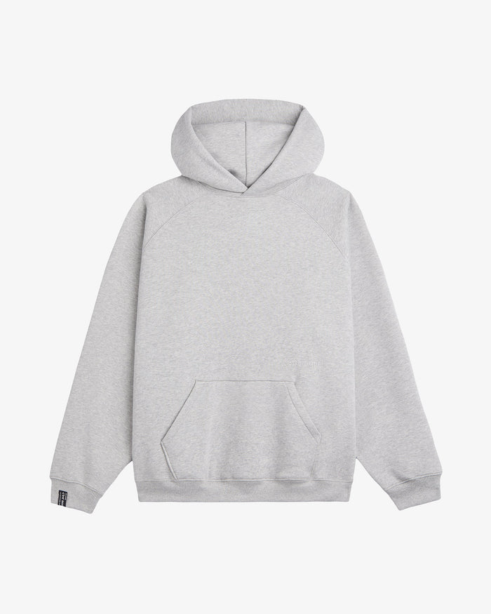 A front view of the Tonal Oversized Hoodie in grey melange, laid flat on a white background. The image highlights the kangaroo pocket, ribbed cuffs and hem, the tone on tone embroidered c'est normal logo on the chest, and the black cuff tag on the right sleeve.