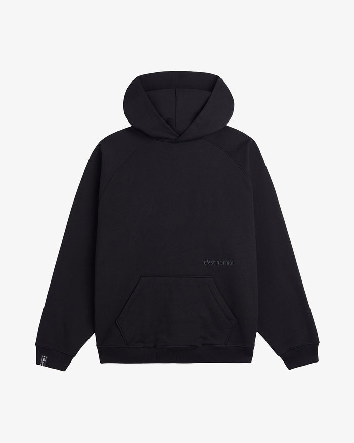 A front view of the Tonal Oversized Hoodie in black, laid flat on a white background. The image highlights the kangaroo front pocket, ribbed cuffs and hem, and the tone-on-tone embroidered c'est normal logo.