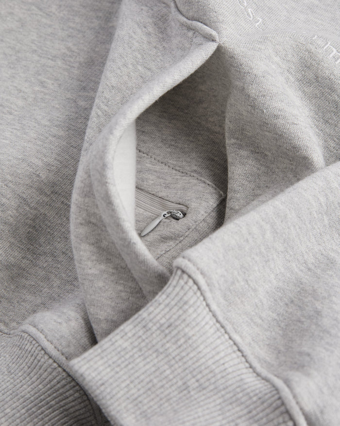 A close-up view of the Tonal Oversized Hoodie in grey melange, highlighting the ribbed hem, the kangaroo front pocket, and the hidden zip pocket inside. The c'est normal embroidered logo is partially visible.