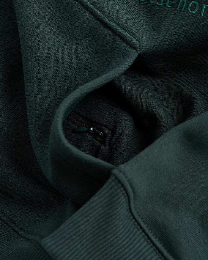 A close-up shot of the Tonal Oversized Hoodie in pine green, highlighting the hidden zip pocket inside the kangaroo front pocket. Part of the tone-on-tone embroidered c'est normal logo is visible at the top.