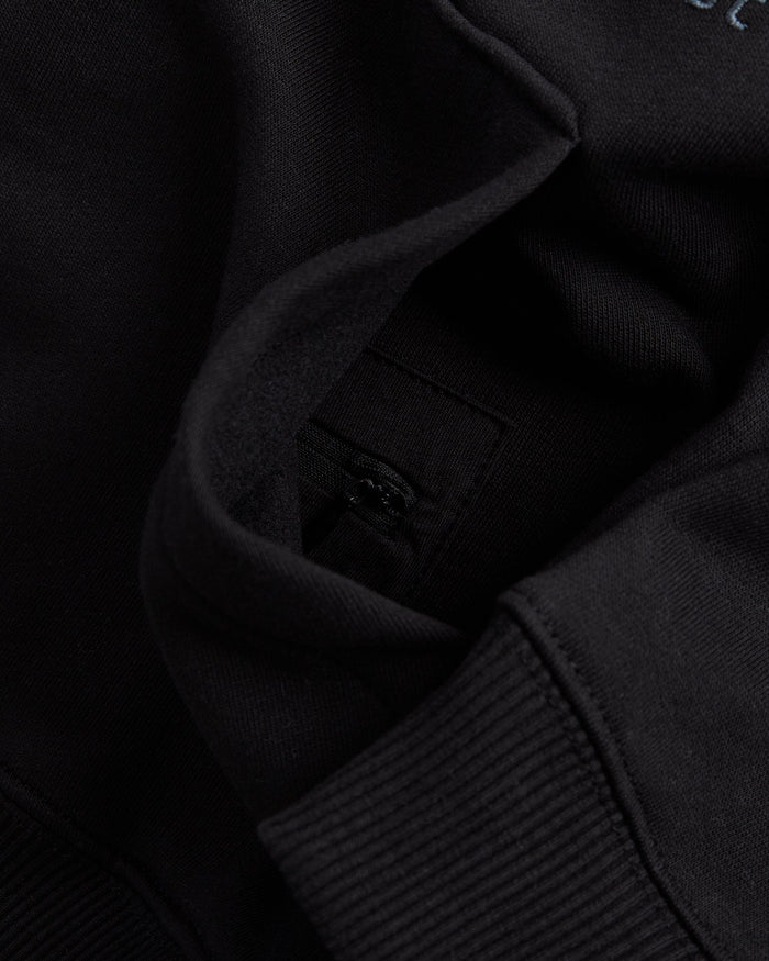 A detailed close-up of the Tonal Oversized Hoodie in black, focusing on the kangaroo front pocket with its hidden zip pocket inside.