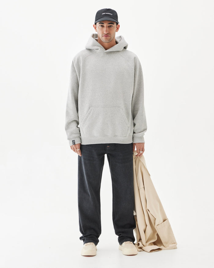 A man wearing the Tonal Oversized Hoodie in Grey Melange, paired with black jeans, a black c'est normal cap, and holding a beige jacket. The image features a full-body shot against a white background.