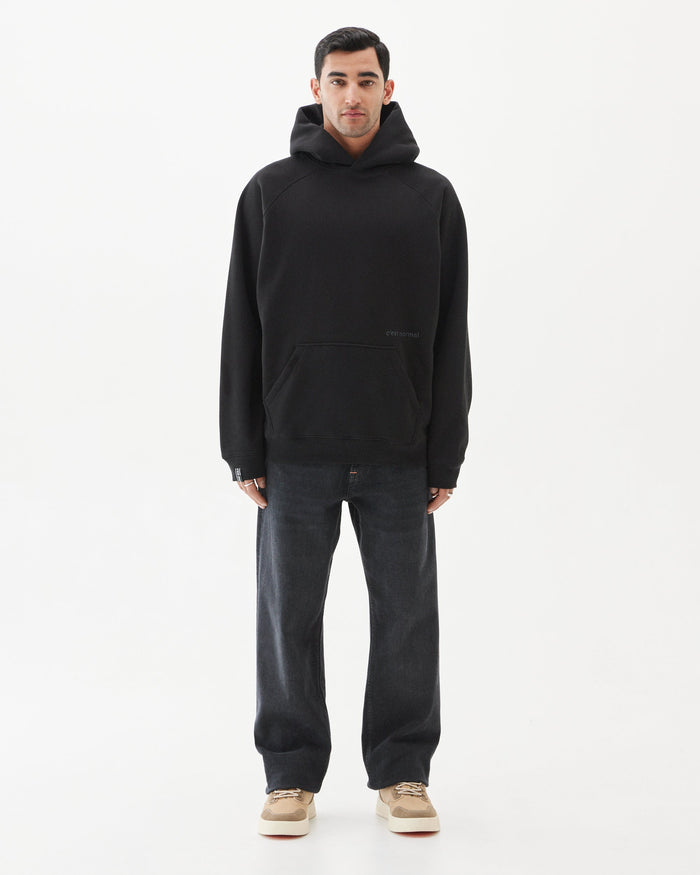 A front view of a man wearing the Tonal Oversized Hoodie in black by c'est normal, styled with dark grey jeans and beige sneakers. The image highlights the oversized fit, kangaroo pocket, and tone on tone embroidered c'est normal logo against a white background.