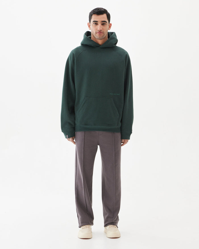 A front view of a man wearing the Tonal Oversized Hoodie in pine, standing against a white background. The image showcases the oversized fit, kangaroo pocket, and tonal embroidered c'est normal logo.