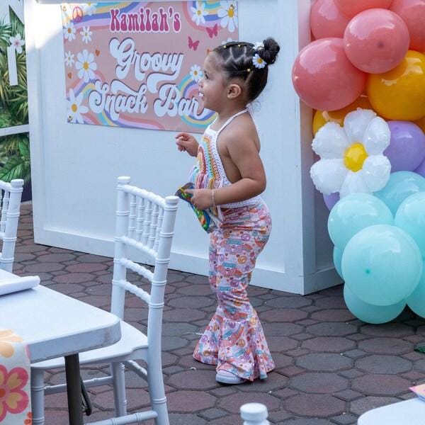 A young girl in a floral dress stands in front of a colorful banner for "Kamilah's Groovy Snack Bar" at a birthday party.