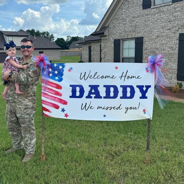 A man in military uniform stands next to a banner that reads "Welcome Home Daddy We missed you!" with a young child in his arms.