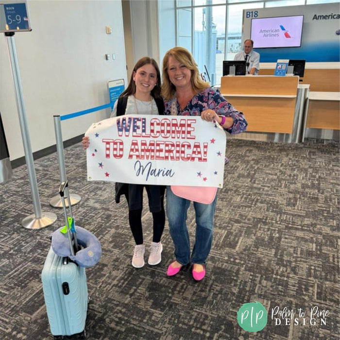 Two women are holding a sign that says "Welcome to America! Maria" in an airport terminal, with a suitcase and a man in a suit visible in the background.