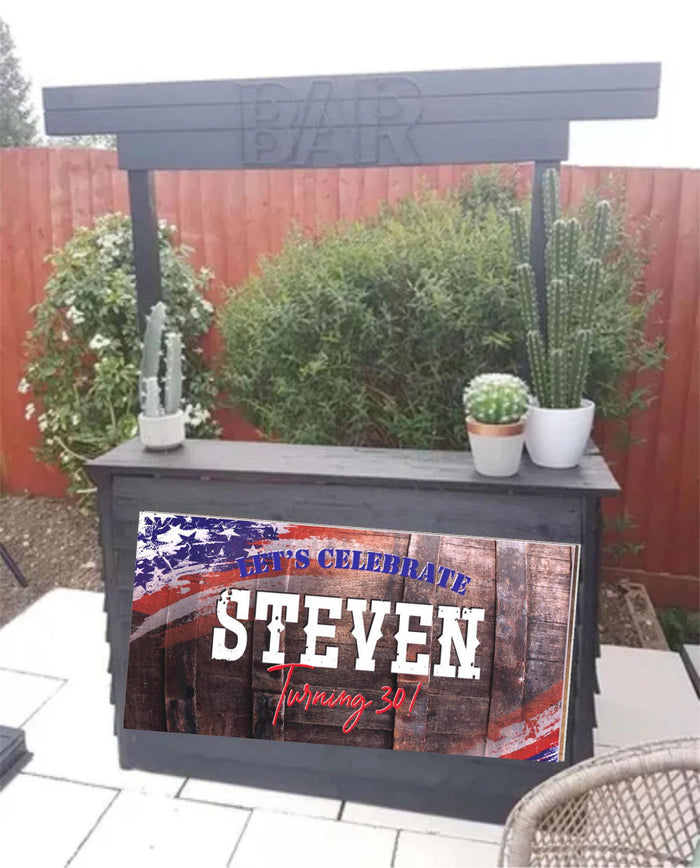 A wooden sign with a red, white, and blue American flag birthday banner design for adult men. The sign says 'Let's Celebrate Steven Turning 30!' The sign is in front of a black wooden bar with a sign that says 'Bar' above it.