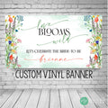 A custom vinyl banner with a watercolor floral design. The banner reads 'love blooms wild' and 'Let's celebrate the bride-to-be' with a personalized name. The banner is green, white, and multicolored flowers with a watercolor effect.