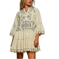 A woman modeling a cream-colored bohemian-style mini dress with blue floral embroidery and lace trim, set against a textured gray studio background.