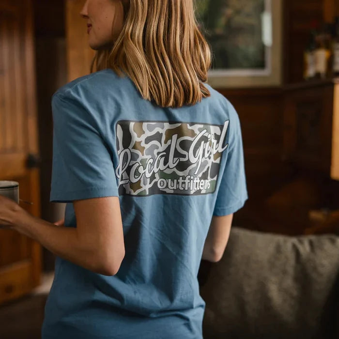 Person wearing a blue t-shirt with a logo and text, holding a cup indoors.